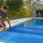 Técnico medindo capa térmica azul parcialmente aberta sobre piscina residencial com casa de máquinas ao fundo