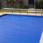 Piscina residencial com capa térmica azul instalada sobre a água e técnico ajustando a borda ao lado do rolo enrolador