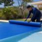 Técnico desenrolando capa térmica azul sobre piscina residencial com enrolador na borda