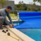Técnico ajustando capa térmica azul com rolo enrolador e trena na borda de piscina residencial