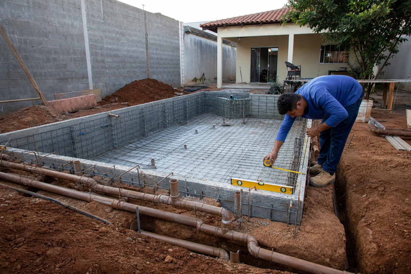Técnico medindo estrutura de piscina em construção com ferragens e tubulação hidráulica aparente