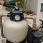 Mão com luva ajustando a válvula seletora de um filtro de areia de piscina com manômetro visível