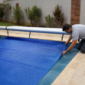 Técnico conferindo a borda de uma capa térmica azul instalada sobre piscina residencial, com enrolador metálico ao lado