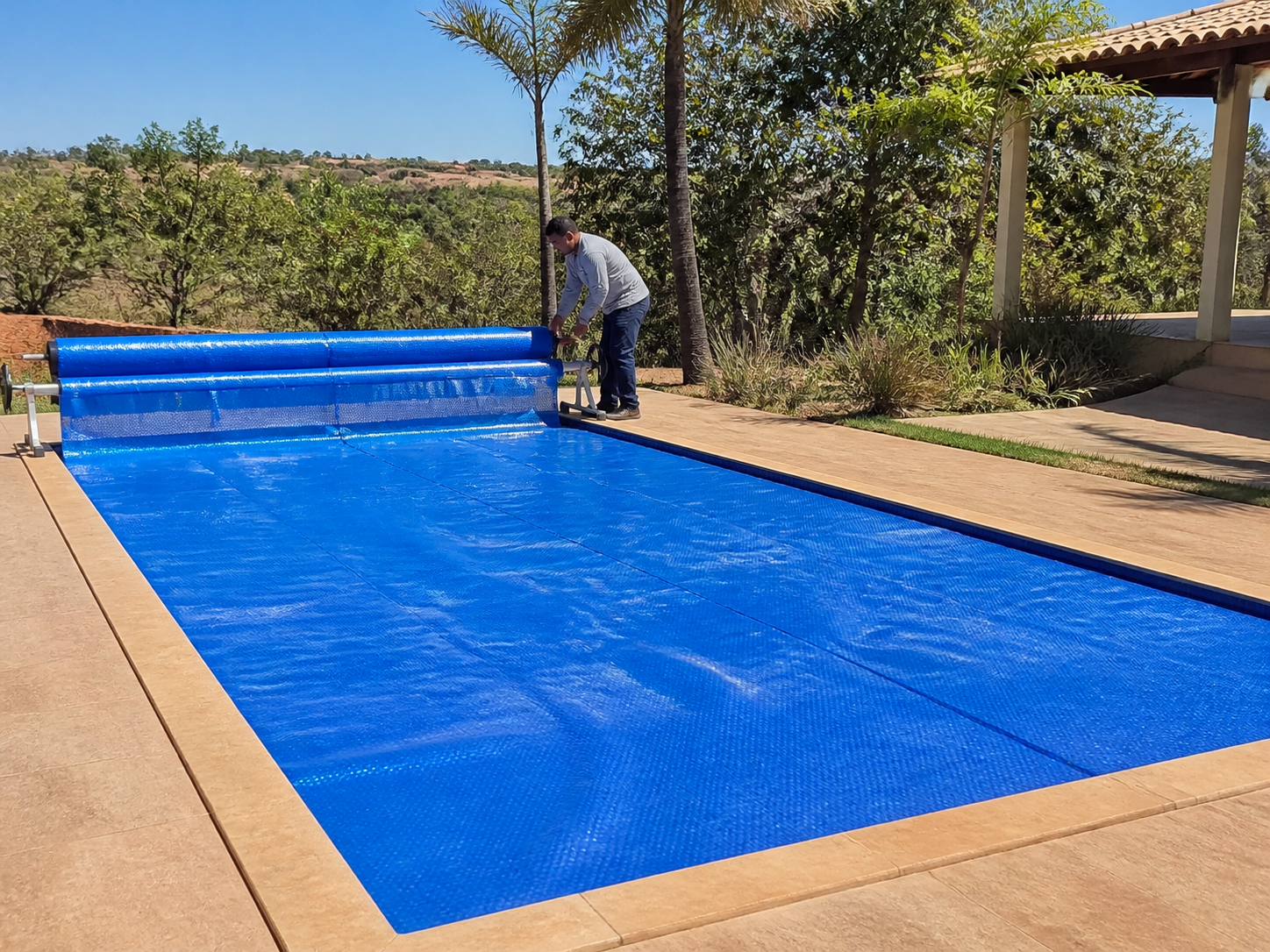 Capa térmica azul cobrindo piscina residencial com técnico conferindo enrolador e encaixe da manta