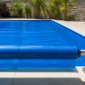 Técnico conferindo a borda de uma capa térmica azul instalada sobre piscina residencial, com enrolador metálico ao lado