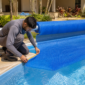 Técnico conferindo a borda de uma capa térmica azul instalada sobre piscina residencial com enrolador metálico