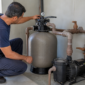 Técnico conferindo filtro de areia, bomba, manômetro e tubulações de PVC na casa de máquinas de uma piscina