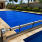 Técnico conferindo a borda de uma capa térmica azul instalada sobre piscina residencial, com enrolador metálico ao lado