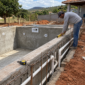 Técnico conferindo o nível de uma piscina em construção com ferragens, concreto e tubulações hidráulicas de PVC